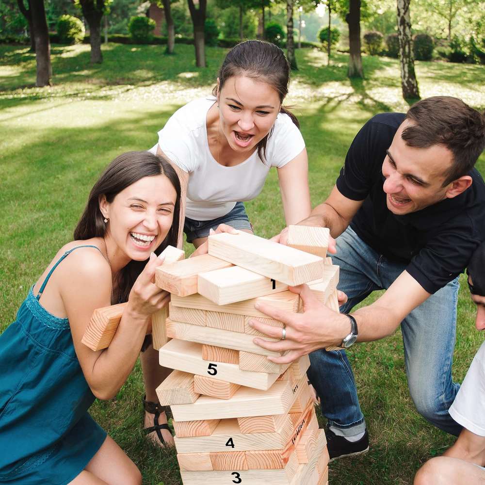 Three adults laughing as they play a giant wooden tumbling tower game on a lawn